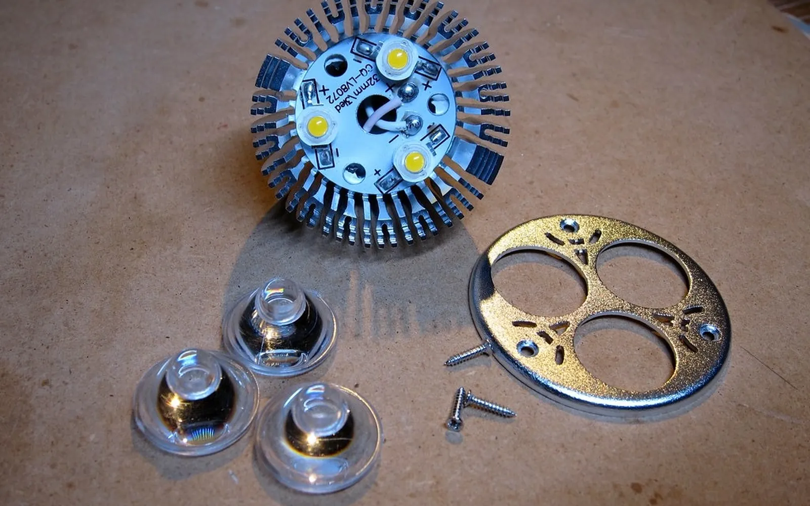 The inside of an LED lamp taken apart, showing three LED chips on a round circuit board, a finned heat sink, clear lenses and screws