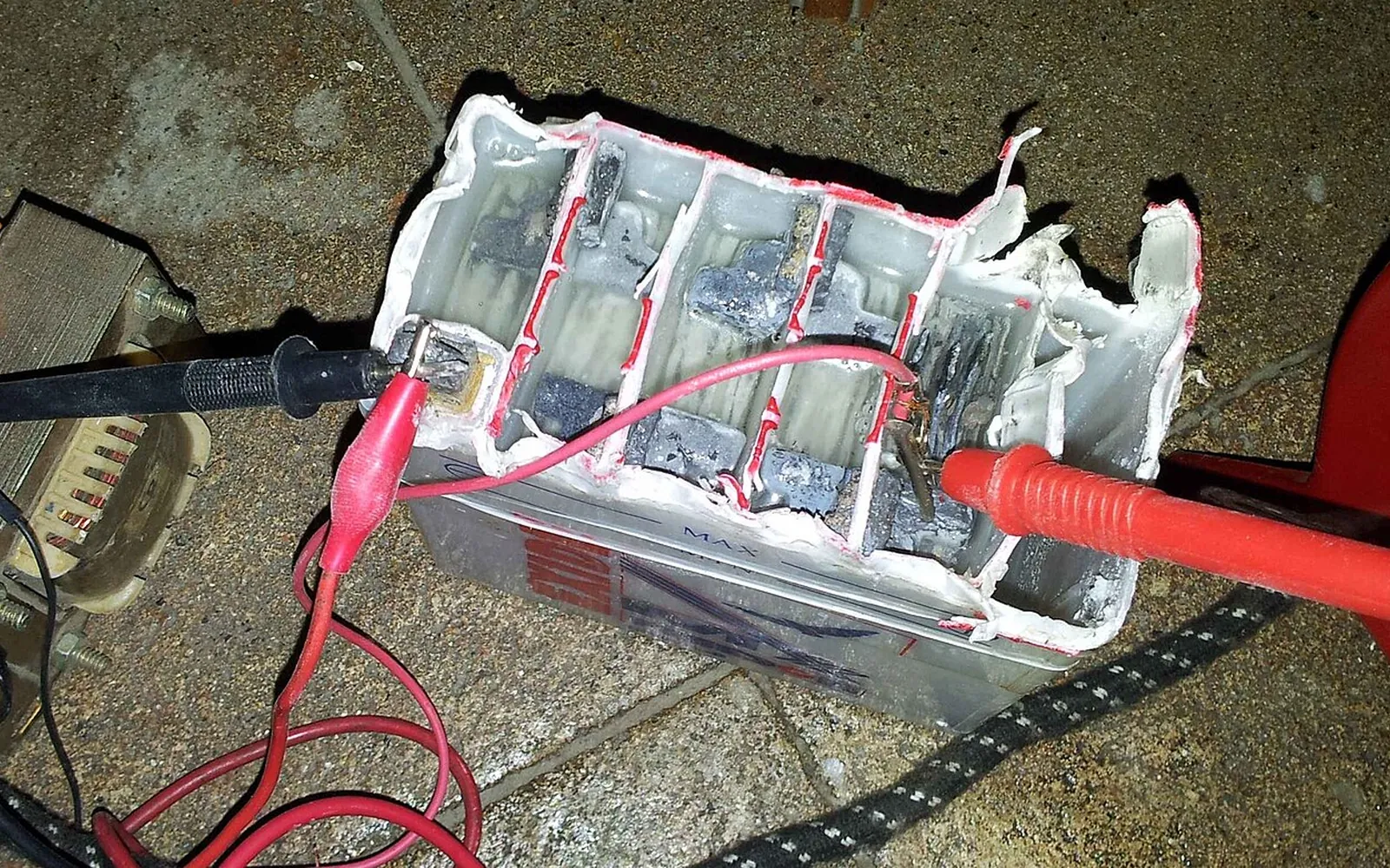 A cut-open lead-acid battery showing its separate cells and grey plates, with multimeter probes clipped to it