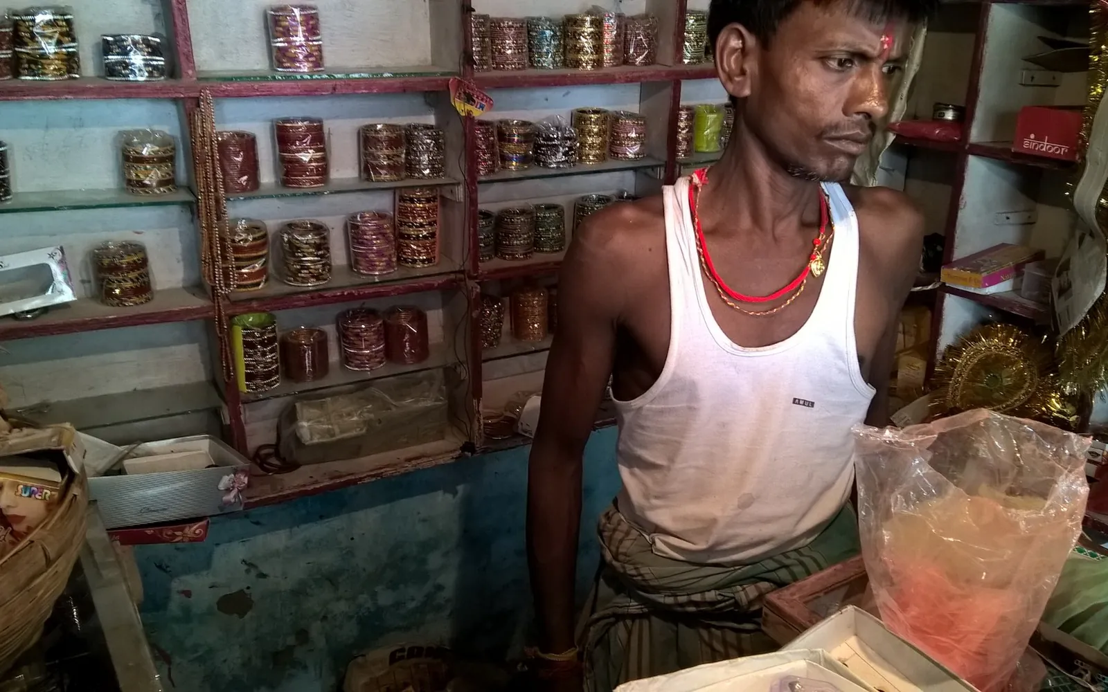 An Indian shopkeeper behind a counter in a small shop lined with shelves