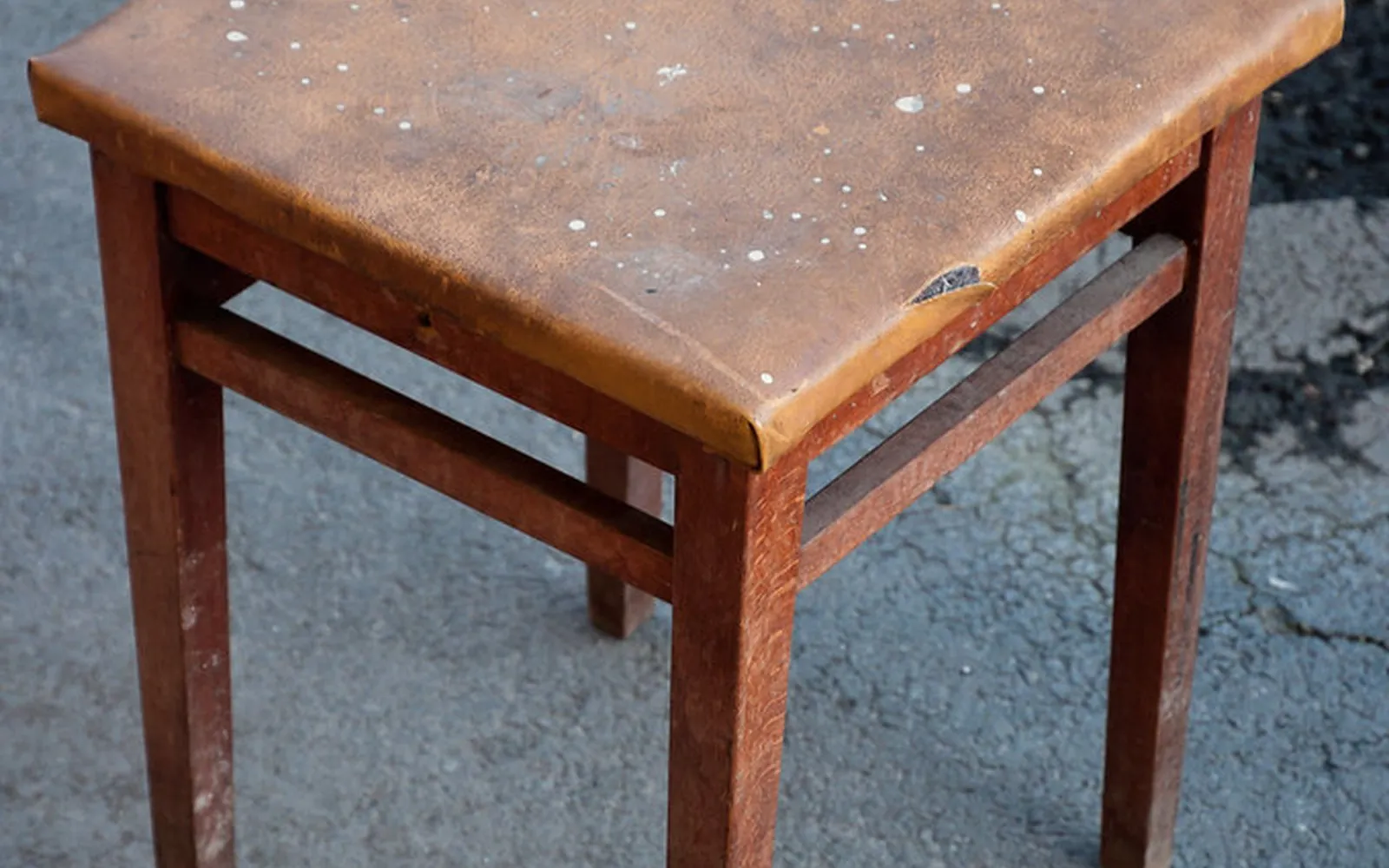 A worn wooden stool with a scuffed brown seat standing on a cracked pavement